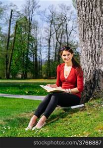 Photo of a beautiful young woman reading a book sitting against a tree in early spring.&#xA;
