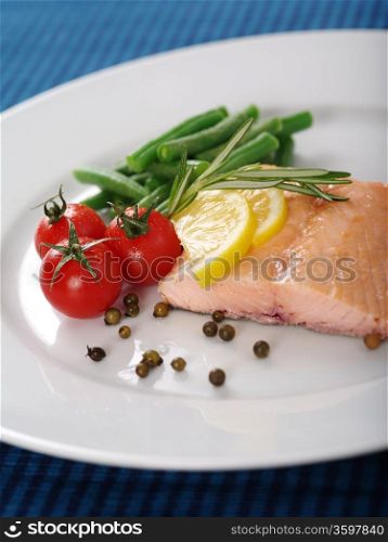 Photo of a cooked salmon steak with rosemary, lemon slices, cherry tomatoes, green beans, and peppercorns on a white plate.