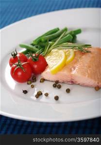 Photo of a cooked salmon steak with rosemary, lemon slices, cherry tomatoes, green beans, and peppercorns on a white plate.