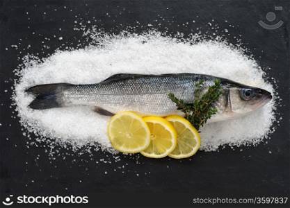 Photo of a full raw fish on a bed of sea salt and on black slate with lemon slices and thyme.