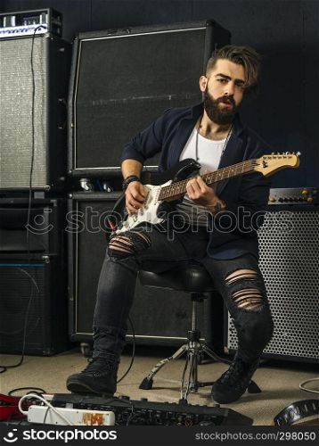 Photo of a man with beard sitting and playing his electric guitar in a recording studio.
