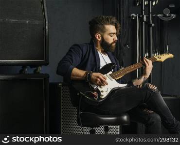 Photo of a man with beard sitting and playing his electric guitar in a recording studio.