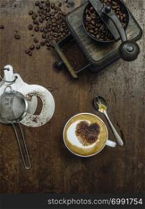 Photo of a messy rustic wooden table of coffee beans, grinder and a cup of coffee with heart-shaped chocolate sprinkle.
