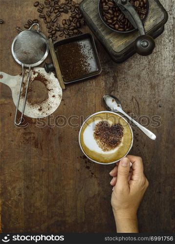 Photo of a messy rustic wooden table of coffee beans, grinder and a woman&rsquo;s hand holding a finished cup.