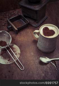 Photo of a messy rustic wooden table of coffee beans, grinder and a cup of coffee with heart-shaped chocolate sprinkle.