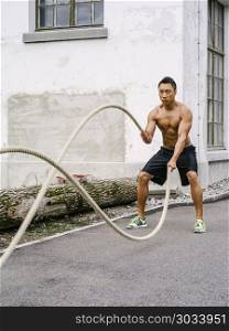 Photo of a muscular Asian man working out with training ropes outside.. Workout outdoors with training ropes