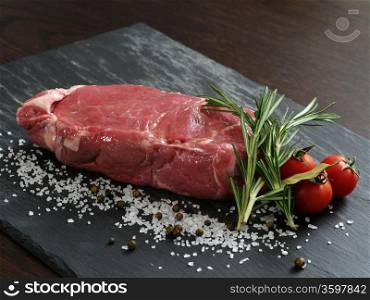 Photo of a raw thick sirloin steak with rosemary, cherry tomatoes, salt and peppercorns on a piece of black slate.
