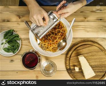 Photo of a woman grating parmesan cheese over a bowl of traditional spaghetti bolognese with a glass of red wine.