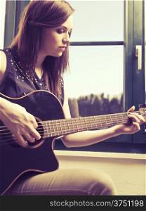 Photo of a woman playing an acoustic guitar by a window. Heavily filtered.
