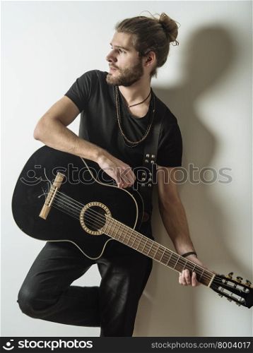 Photo of a young attractive man with long hair and beard leaning against the wall holding an acoustic guitar.&#xA;