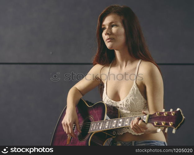 Photo of a young beautiful redhead woman sitting playing an acoustic guitar.