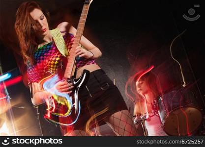 Photo of a young beautiful woman playing guitar with drummer in the background. Motion blur and light trails from slow shutter speed and strobes. Space for text.