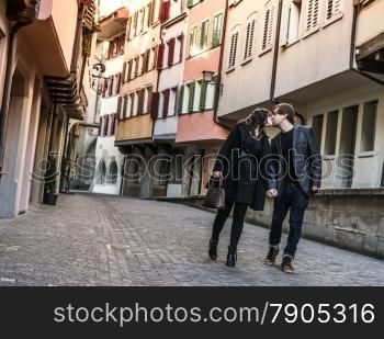 Photo of a young couple walking and kissing in an old town in Europe.