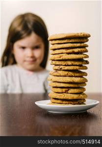 Photo of a young girl staring at a tall pile of chocolate chip cookies. Focus on the cookies.