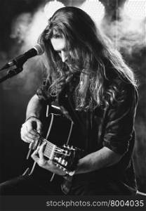 Photo of a young man with long hair and a beard singing and playing an acoustic guitar on stage with lights and concert atmosphere.&#xA;
