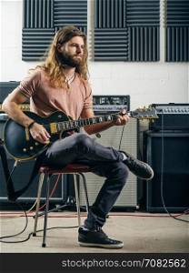 Photo of a young man with long hair and beard playing electric guitar in a recording studio.