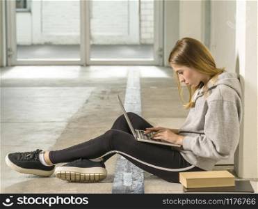 Photo of a young woman student sitting in a hallway with a laptop and books.