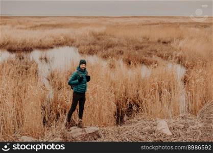 Photo of active male dressed in fashionable clothes, stands on stone against near small lake and yellow autumn field, carries backpack, copy space aside for your advertising content or information