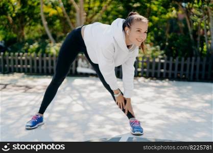 Photo of active pleased woman smiles gladfully concentrated into distance leans to leg dressed in sportswear trains outdoors being motivated listens music in wireless earphones does aerobic exercises