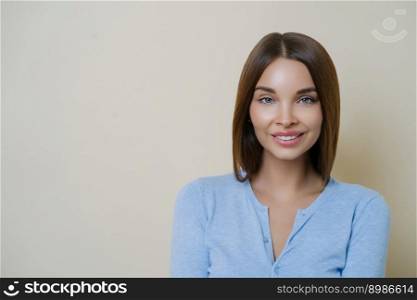 Photo of adorable brunette woman has natural makeup and healthy perfect skin, smiles tenderly, shows white teeth, spends free time on caring about herself, wears casual blue jumper, poses indoor