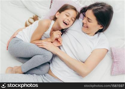 Photo of adorable small child and her mother have fun together in bed, tickle each other, smile joyfully, play after good sleep, have good relationships. People, family, motherhood and childhood