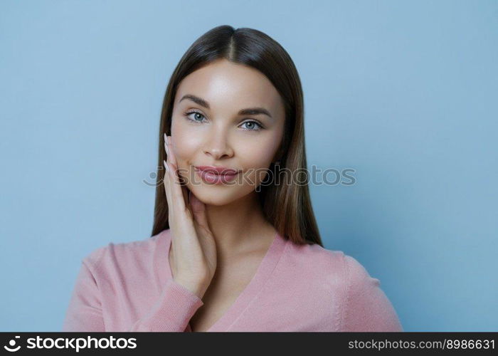 Photo of adorable young millennial female touches cheek gently, enjoys softness of skin after applying beauty mask or face cream, gazes at camera, isolated over blue background. Feminity concept