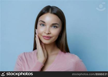 Photo of adorable young millennial female touches cheek gently, enjoys softness of skin after applying beauty mask or face cream, gazes at camera, isolated over blue background. Feminity concept