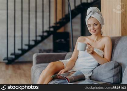 Photo of adorable young woman has makeup and healthy skin, holds mug of coffee, wrapped in bath towel, reads magazine and feels relaxed sitting on sofa, spends free time at home. Feeling comfort