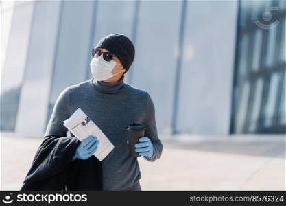 Photo of adult man walks at street, wears protective mask and rubber gloves as protection against infectious disease, drinks takeaway coffee, bought newspaper to read news, concentrated aside