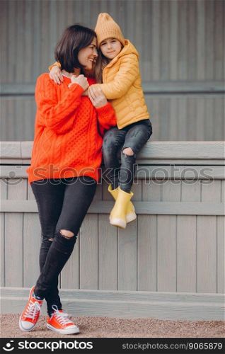 Photo of affectionate mother spends free time together with daughter, pose closely to each other, dressed in black ripped jeans and casual clothes, have happy expressions. Parenthood concept