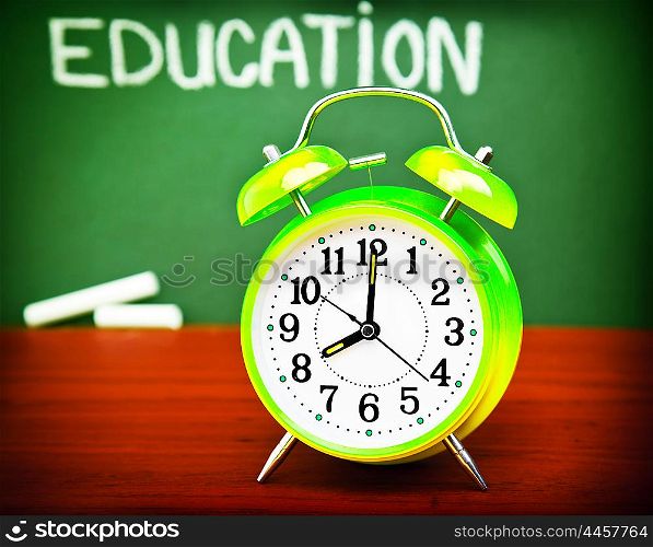 "Photo of alarm clock on wooden desk in classroom, handwriting word "education" on green blackboard, autumn season, yellow watch in school class, back to school, knowledge concept&#xA;"