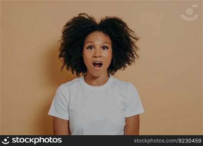 Photo of amazed stunned dark skinned female looking at camera with opened mouth, shocked african woman in white tshirt with surprised face expression posing against beige background. Amazed stunned dark skinned female looking at camera with opened mouth isolated over beige wall