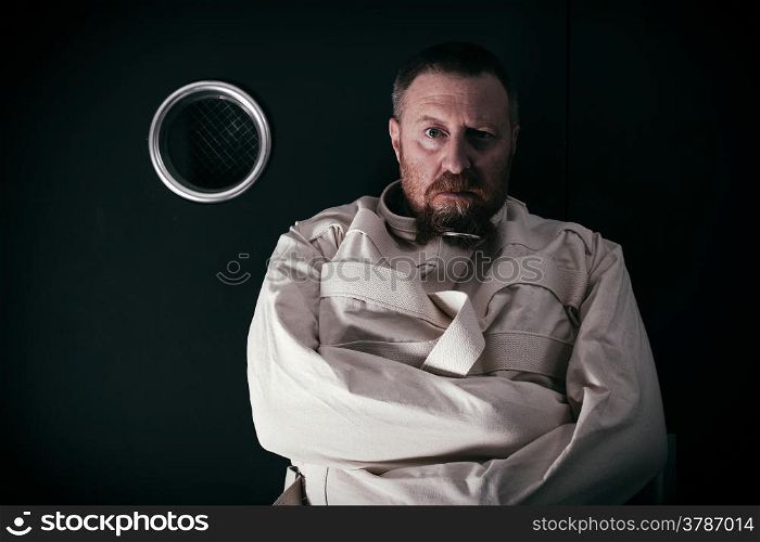 Photo of an insane man in his forties wearing a straitjacket standing in a cell of an asylum with the light from the hallway streaming in.