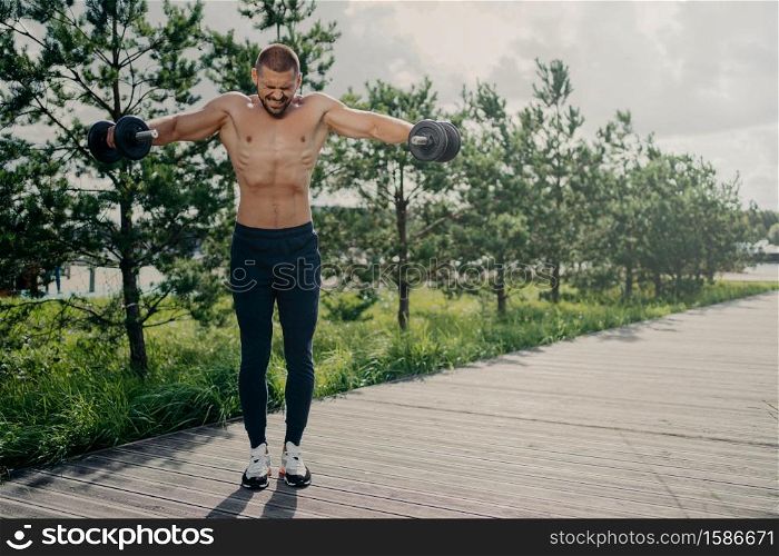 Photo of athletic strong fitness man works out with barbells, demonstrates power and energy, leads active lifestyle, shows strength, motivation, does biceps lifting, has physical workout outdoor