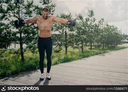 Photo of athletic strong fitness man works out with barbells, demonstrates power and energy, leads active lifestyle, shows strength, motivation, does biceps lifting, has physical workout outdoor
