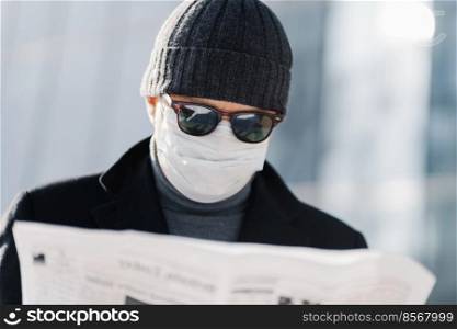 Photo of attentive man wears blak sunglasses, hat and medical mask on face during quarantine and coronavirus outbreak, reads press, finds out news from newspaper, stands against blurred background