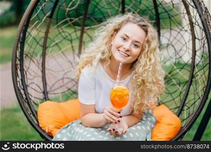 Photo of attractive beautiful curly light haired female with cheerful expression, recreats outdoor in hanging chair, drinks cocktail enjoys sunny day during summer. People, rest and lifestyle concept.
