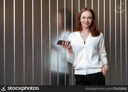 Photo of attractive brown haired businesswoman wears formal black and white clothes, uses app on smartphone, has make up and manicure, keeps hand in pocket, stands against wall with blank space