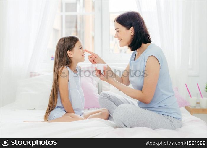 Photo of attractive brunette young female holds bottle of cream, applies it on daughter`s skin, have positive expressions after healthy sleep, pose together on comfortable bed, dressed casually