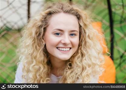 Photo of attractive curly blonde female with shining smile, shows white even teeth, being in high spirit, enjoys good rest outdoor, talks with friend. European woman poses in hanging chair outside