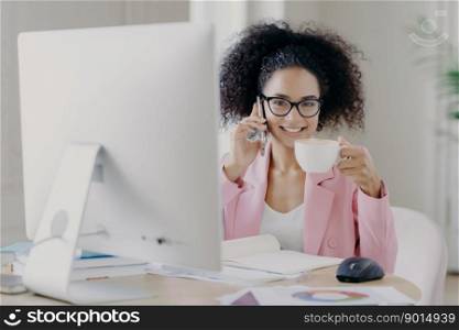 Photo of attractive curly haired elegant woman drinks aromatic coffee, poses at desktop with modern computer needed for work, calls friend during break, discusses latest news, has toothy smile