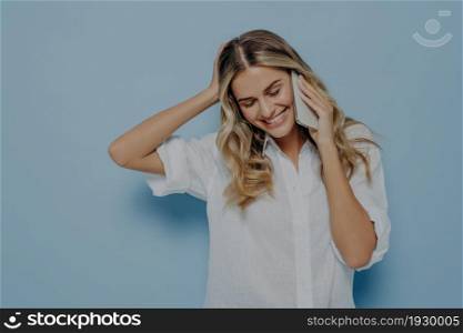 Photo of attractive cute young blonde european woman wearing casual white shirt talking on mobile phone, smiling tenderly and bashfully looking down, isolated over blue background with copy space. Attractive young blonde girl talking on mobile phone, smiling tenderly and bashfully looking down
