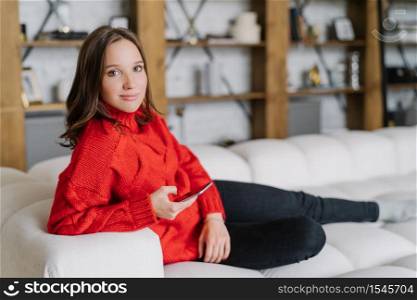 Photo of attractive female model in knitted red sweater, messages online at home, poses on couch in living room, uses modern cell phone and wireless internet connection, looks with appealing look