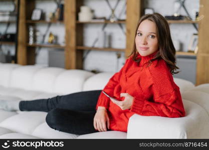 Photo of attractive female model in knitted red sweater, messages online at home, poses on couch in living room, uses modern cell phone and wireless internet connection, looks with appealing look