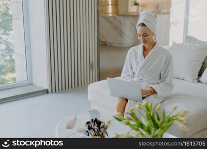 Photo of attractive female model wears bathrobe and towel types on laptop computer works freelance sits on comfortable sofa enjoys domestic atmosphere poses against modern apartment interior