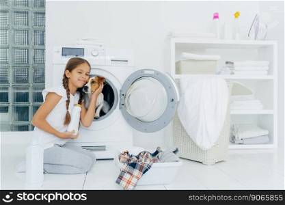 Photo of attractive girl petting pedigree dog in washing machine, holds washing detergent, going to load washer, busy with laundry and domestic chores, washes clothes at home, poses indoors.