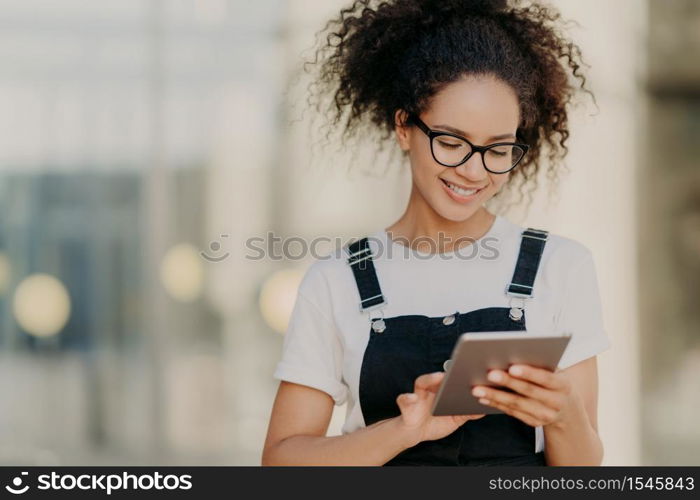 Photo of attractive millennial girl with crisp hair, holds digital tablet, surfes social networks, wears optical glasses, casual t shirt and sarafan, connected to wireless internet, types message