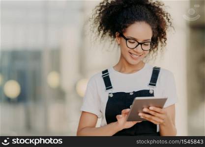 Photo of attractive millennial girl with crisp hair, holds digital tablet, surfes social networks, wears optical glasses, casual t shirt and sarafan, connected to wireless internet, types message