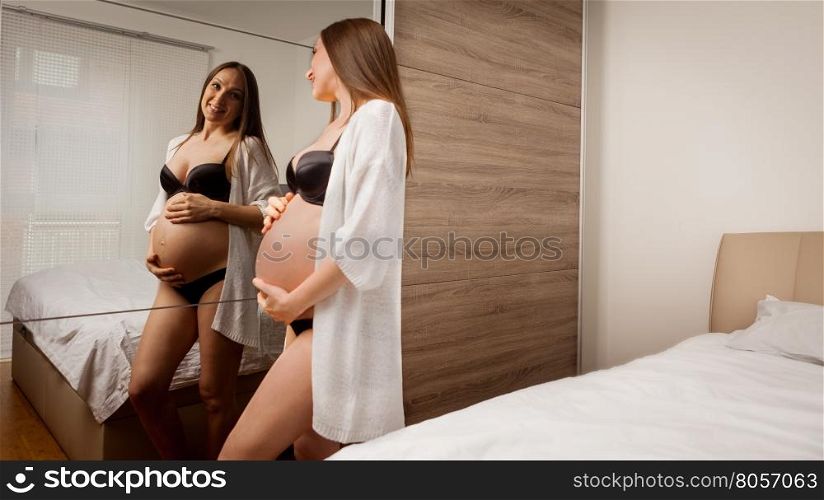 Photo of attractive pregnant woman in lingerie posing in front of her bed room mirror