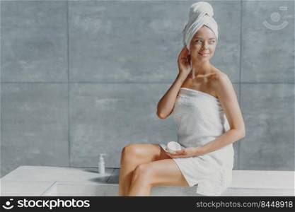 Photo of attractive thoughtful young woman looks away with dreamy expression, puts cream on body after taking bath, has clean fresh skin, poses with bare shoulders, enjoys taking spa procedures